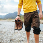 un uomo con delle scarpe in mano cammina a piedi nudi su una riva di un fiume
